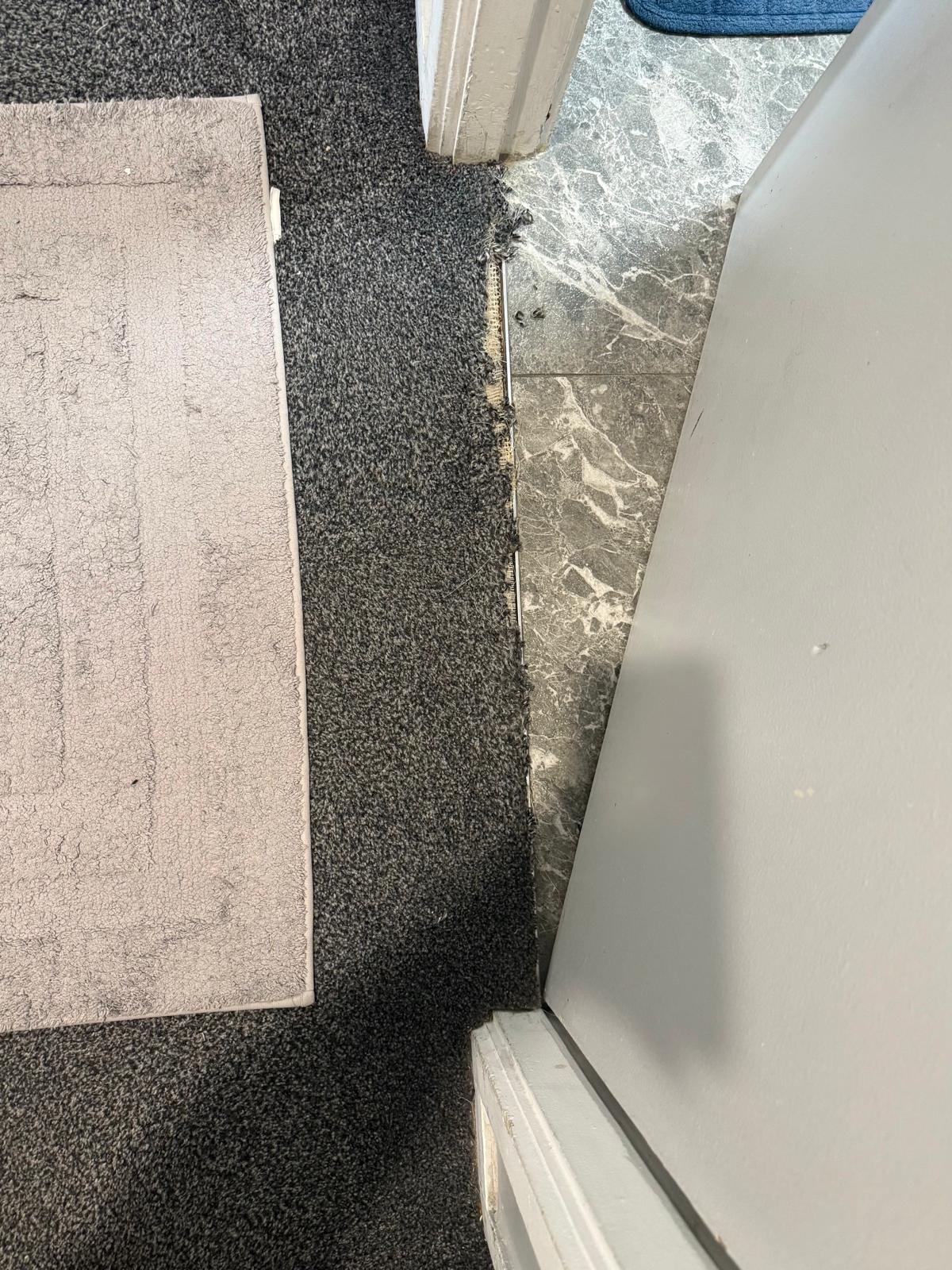 Charcoal carpet frayed and pulling away from the gripper at a doorway beside grey marble tile